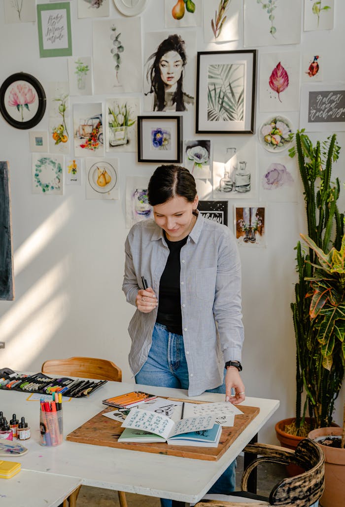 Home Cheerful young female designer in jeans standing near wall with pictures and table with stationery while choosing drafts with letter design in daylight